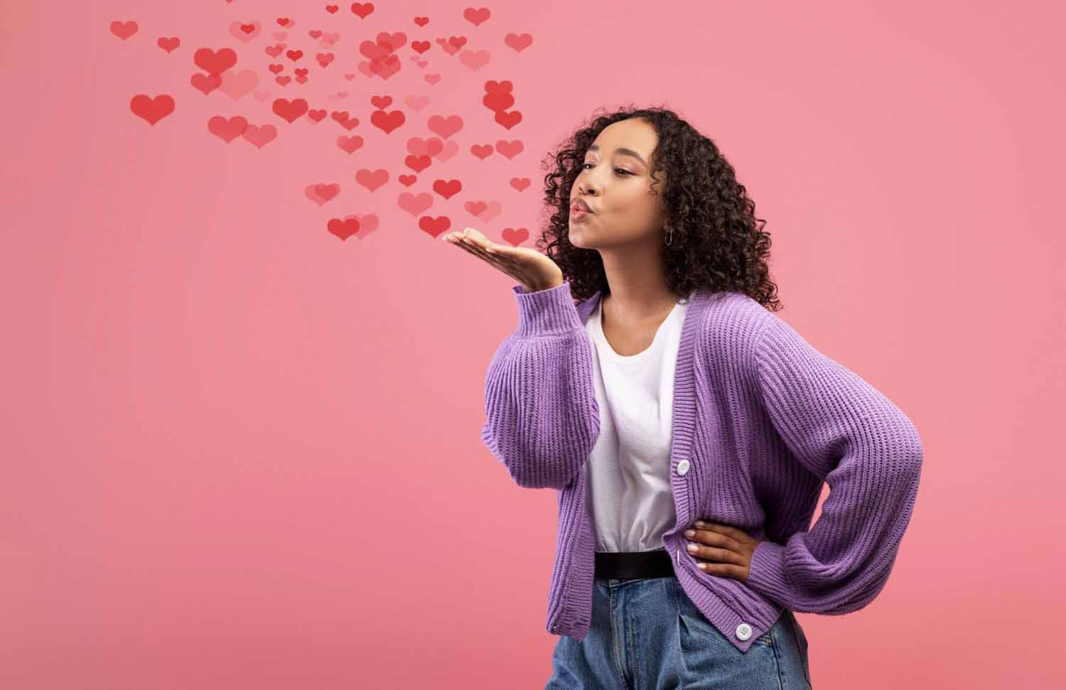 Alt: Young woman blowing air kiss with flying red hearts over pink studio background