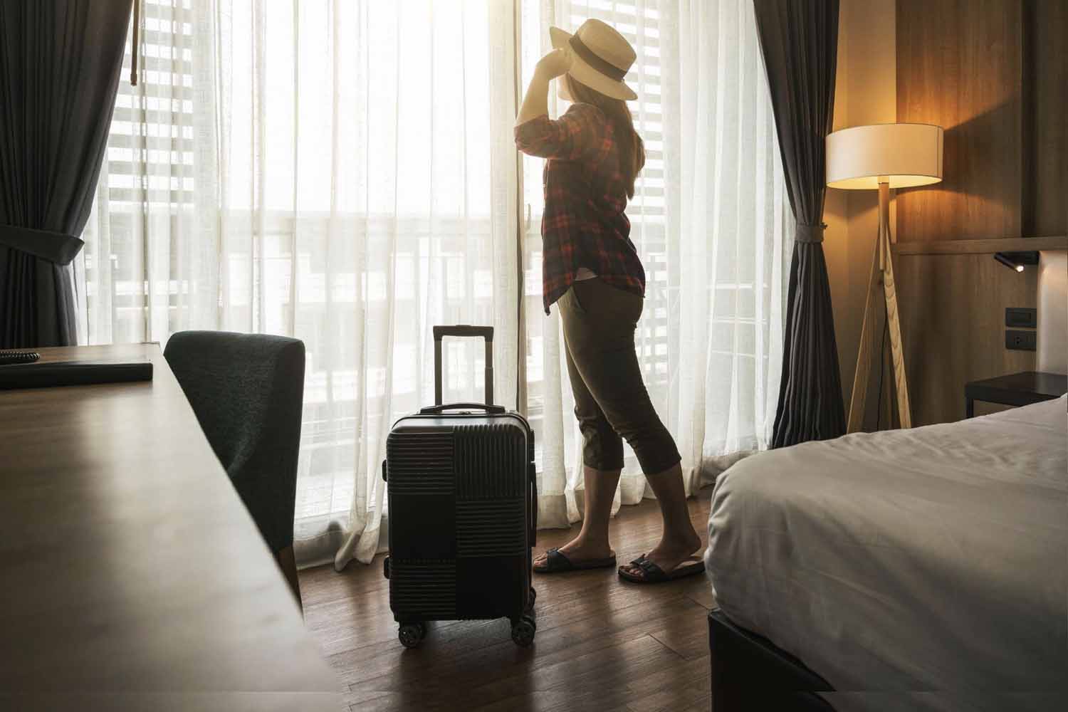 Woman in her hotel room, looking out the window. Suitcase in foreground.