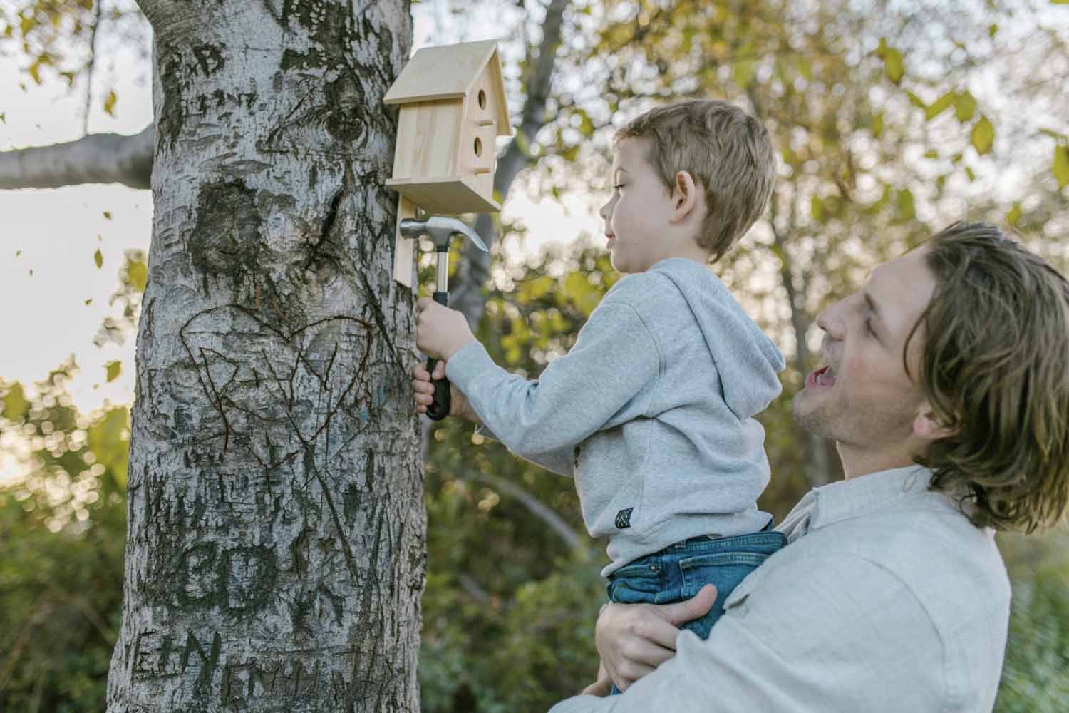 Man holding his son up to a tree while the son uses a hammer to secure the new nesting box they’ve just made