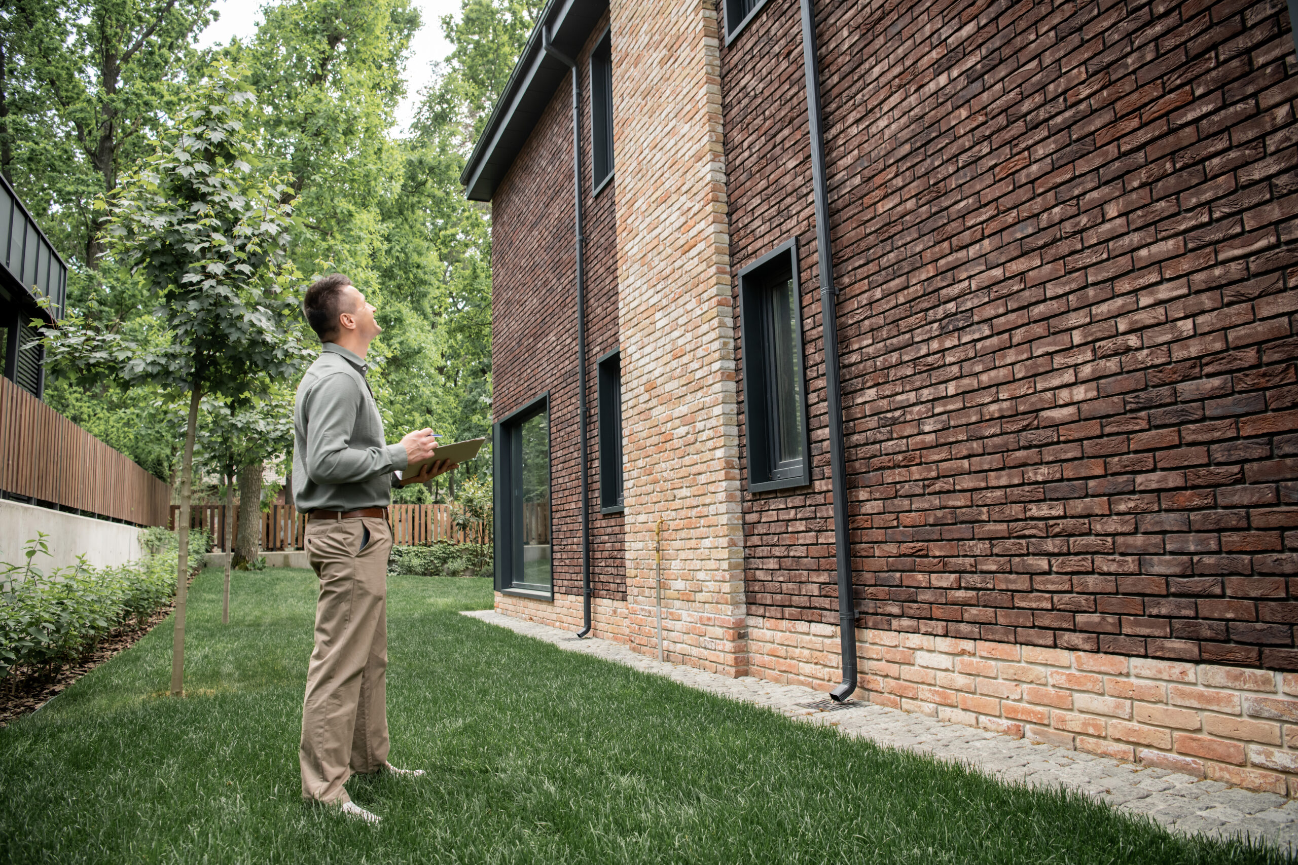 A worker in casual clothing standing in a yard observing a home and conducting an outdoor pest inspection.